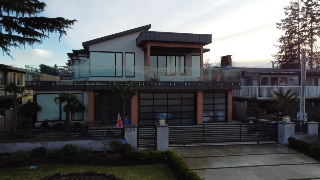 Modern two-story house at 4320 River Road with large windows, a balcony with glass railing, and surrounding greenery under a clear sky.