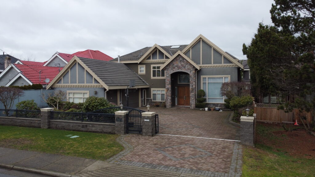 Aerial view of a large, modern two-story house located on Lucas Road with a gray and stone facade, red and gray roofs, and a paved driveway, surrounded by other houses and trees on an