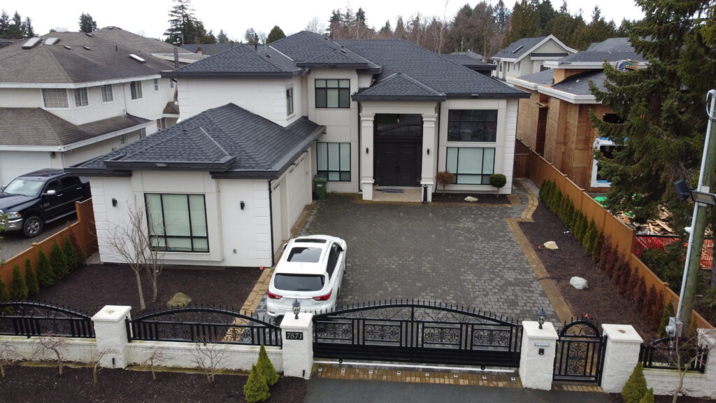 Aerial view of a large modern house at 7571 Bridge Street with a gray roof, featuring a cobblestone driveway, white SUV, and black gate, surrounded by other suburban homes and trees