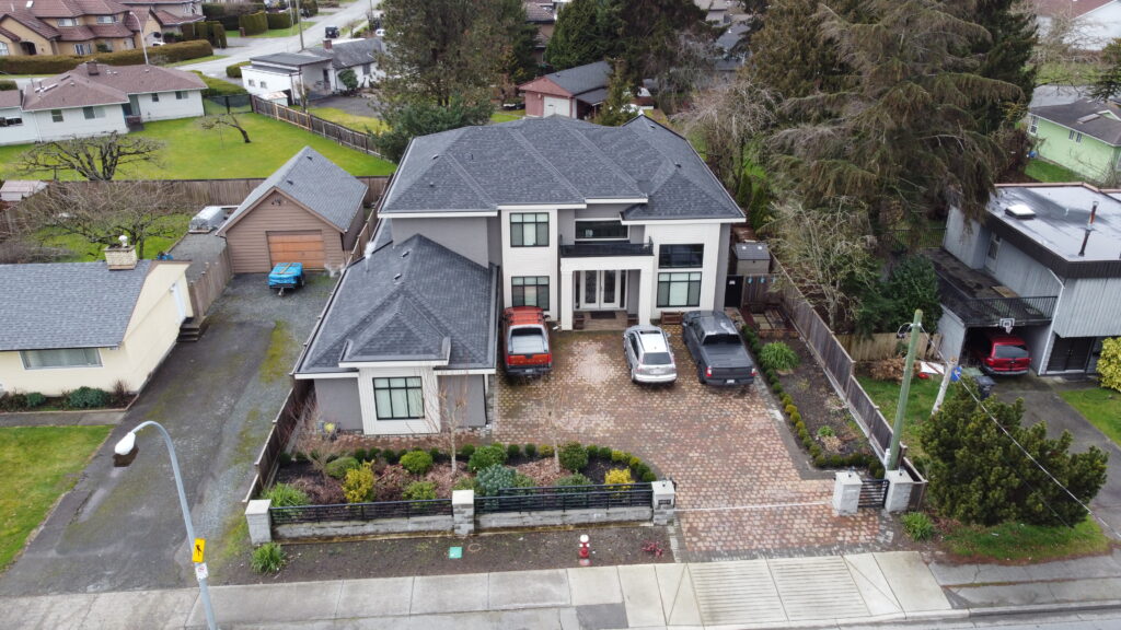 Aerial view of a large, two-story property listing on No. 4 Road with a gray roof, surrounded by smaller homes, featuring a driveway with three cars and landscaped front yard.