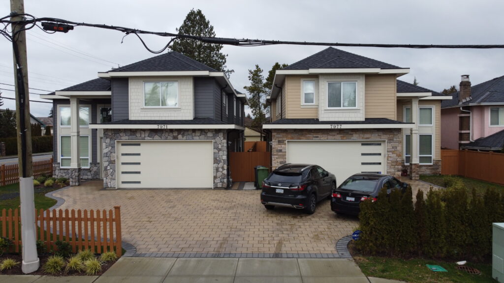 Two modern two-story houses on Bowcock Road with gray siding and stone accents feature front-facing double garages and parked cars in the driveways.
