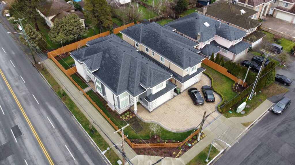 Aerial view of a large gray house with a driveway, parked cars, and landscaped front yard, located beside Bowcock Rd2 in a residential development area.