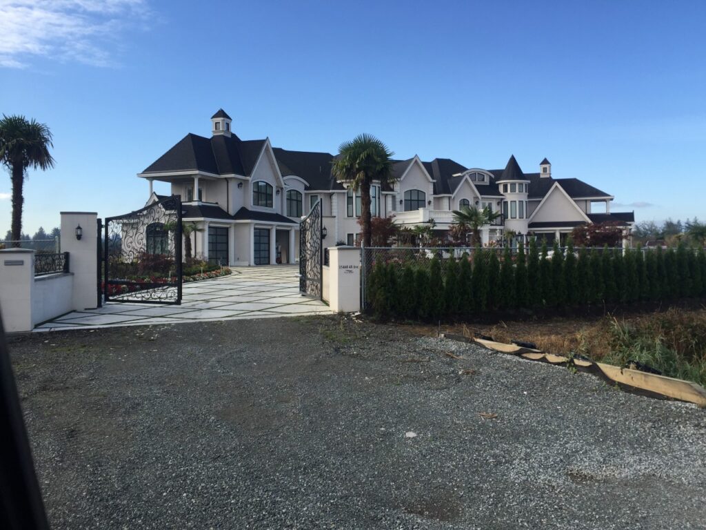 A large, luxurious mansion in South Surrey with multiple gables and a landscaped front yard, viewed from a gravel driveway.